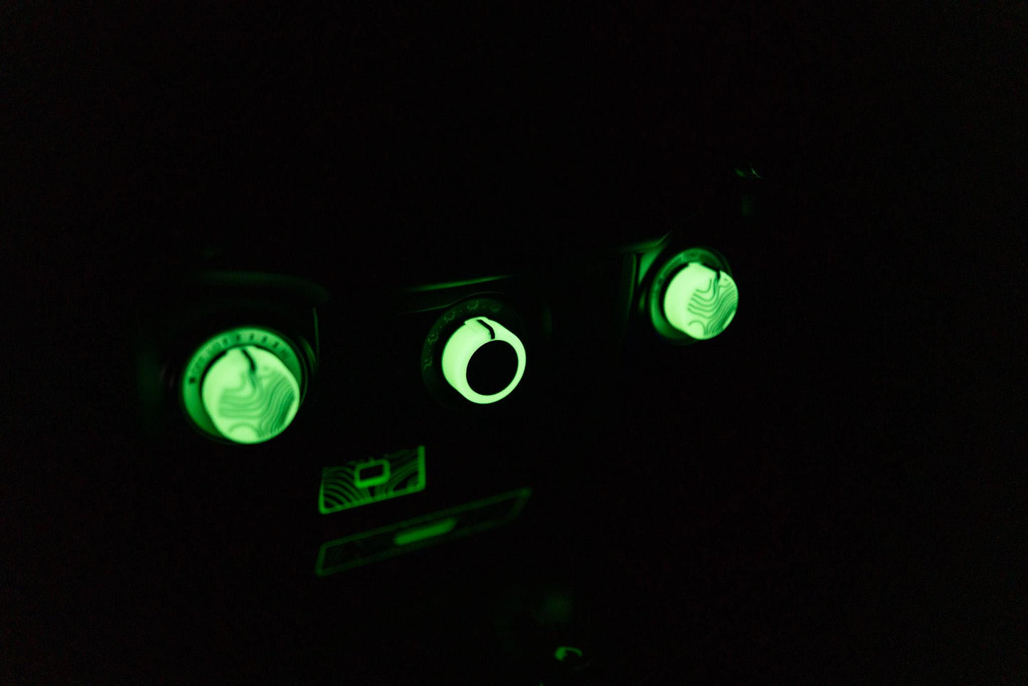 May include: Close-up of a dark control panel with three illuminated green dials and a rectangular display. The dials have a textured, patterned surface. The display and dials glow brightly against the black background.