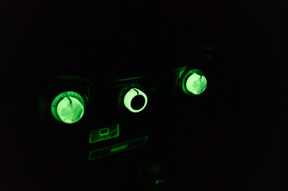 May include: Close-up of a dark control panel with three illuminated green dials and a rectangular display. The dials have a textured, patterned surface. The display and dials glow brightly against the black background.