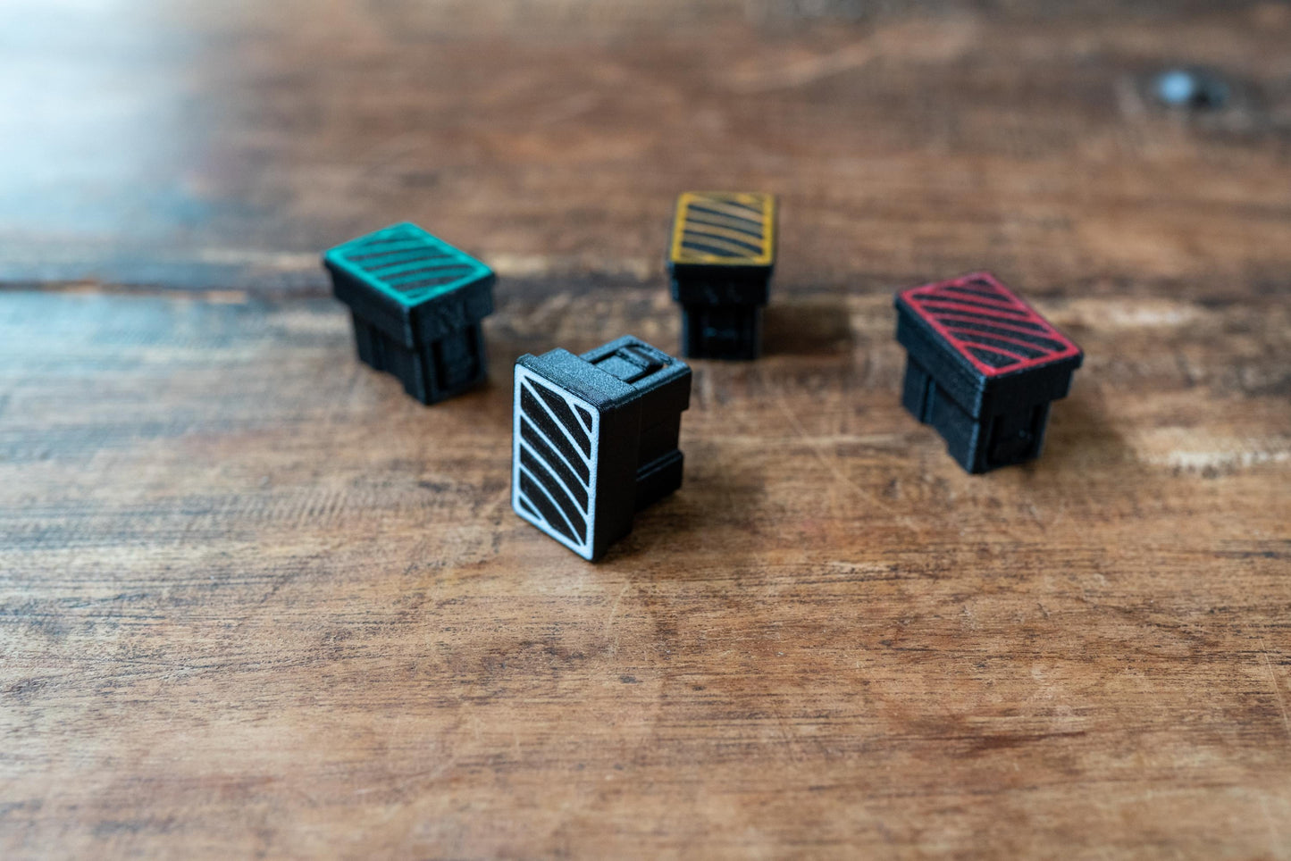 May include: Four small, black, rectangular objects with colored tops and white or colored line designs. The tops are green, yellow, red, and black with white lines. The objects are on a wooden surface.