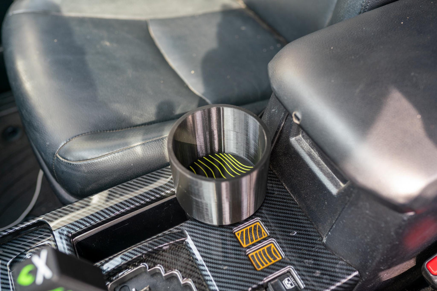 May include: A silver car cup holder with a black and yellow wavy design on the bottom. The cup holder is cylindrical and sits in a car's center console. The interior of the car is black.