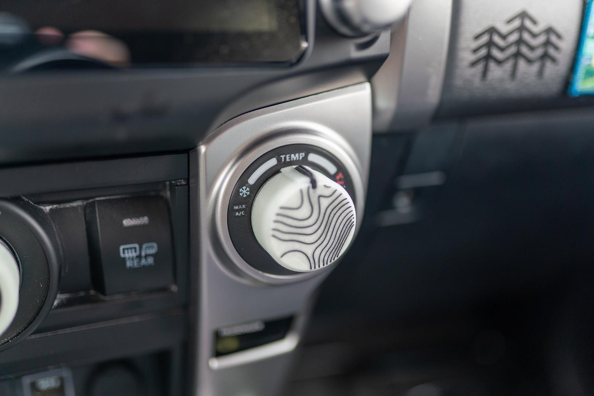 May include: Close-up of a car's dashboard featuring a climate control knob with a white face and black topographic lines. The knob is labeled "TEMP" and has settings for air conditioning. The dashboard is silver and black.