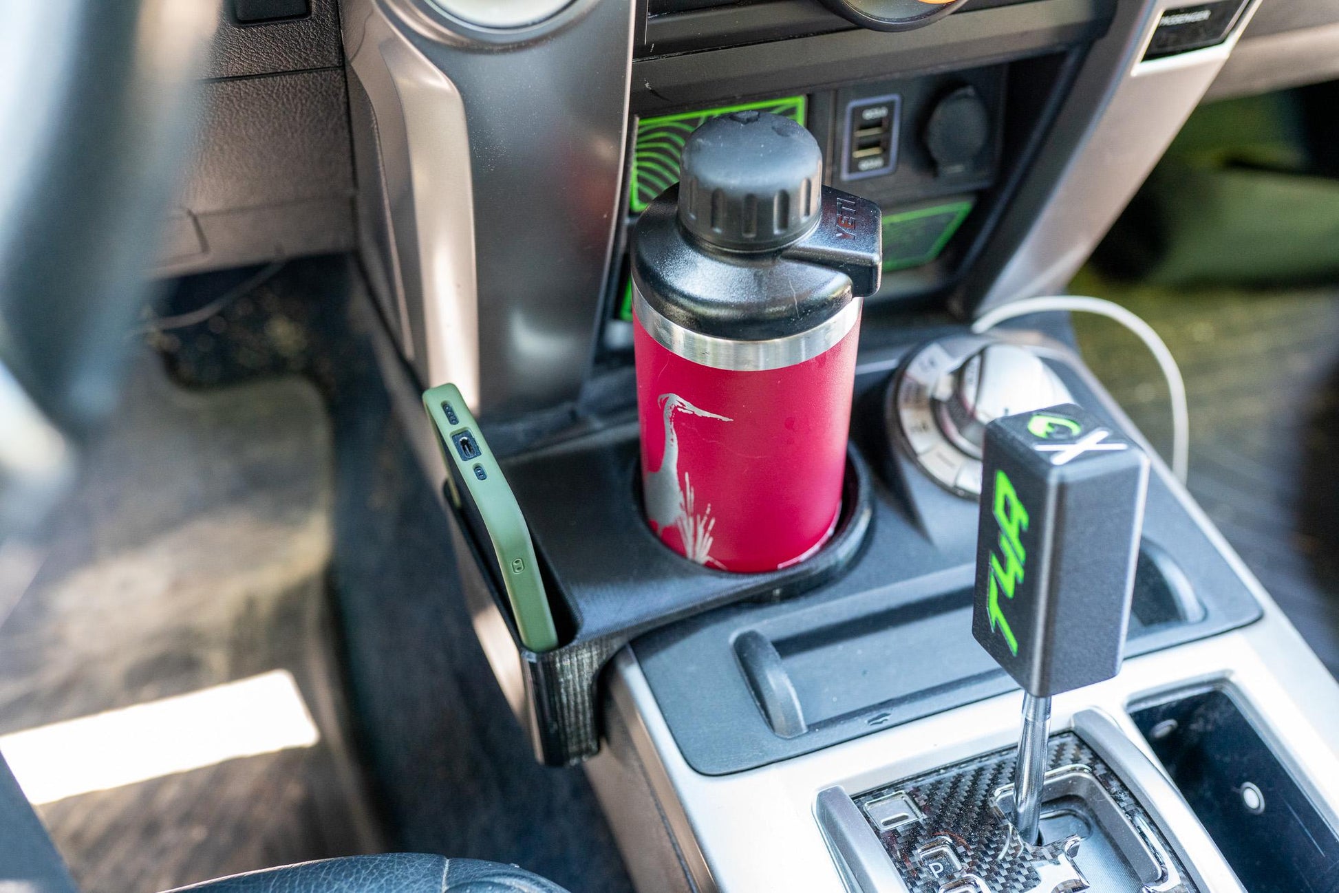 May include: A red insulated water bottle with a heron graphic sits in a car cup holder. A green phone case is in a side slot. The car's interior features a black gear shift and a black cup holder.