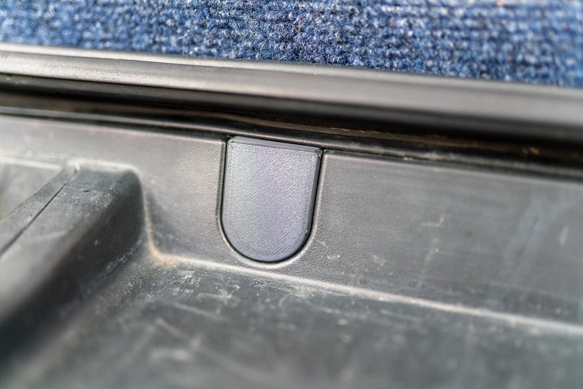 May include: Close-up of a black plastic car part with a U-shaped indentation. The part appears to be a latch or a handle, with a textured surface. The background is a blue carpet.