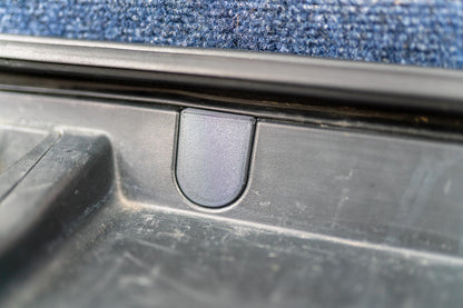 May include: Close-up of a black plastic car part with a U-shaped indentation. The part appears to be a latch or a handle, with a textured surface. The background is a blue carpet.