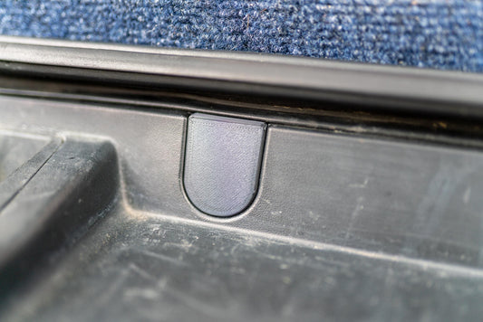 May include: Close-up of a black plastic car part with a U-shaped indentation. The part appears to be a latch or a handle, with a textured surface. The background is a blue carpet.