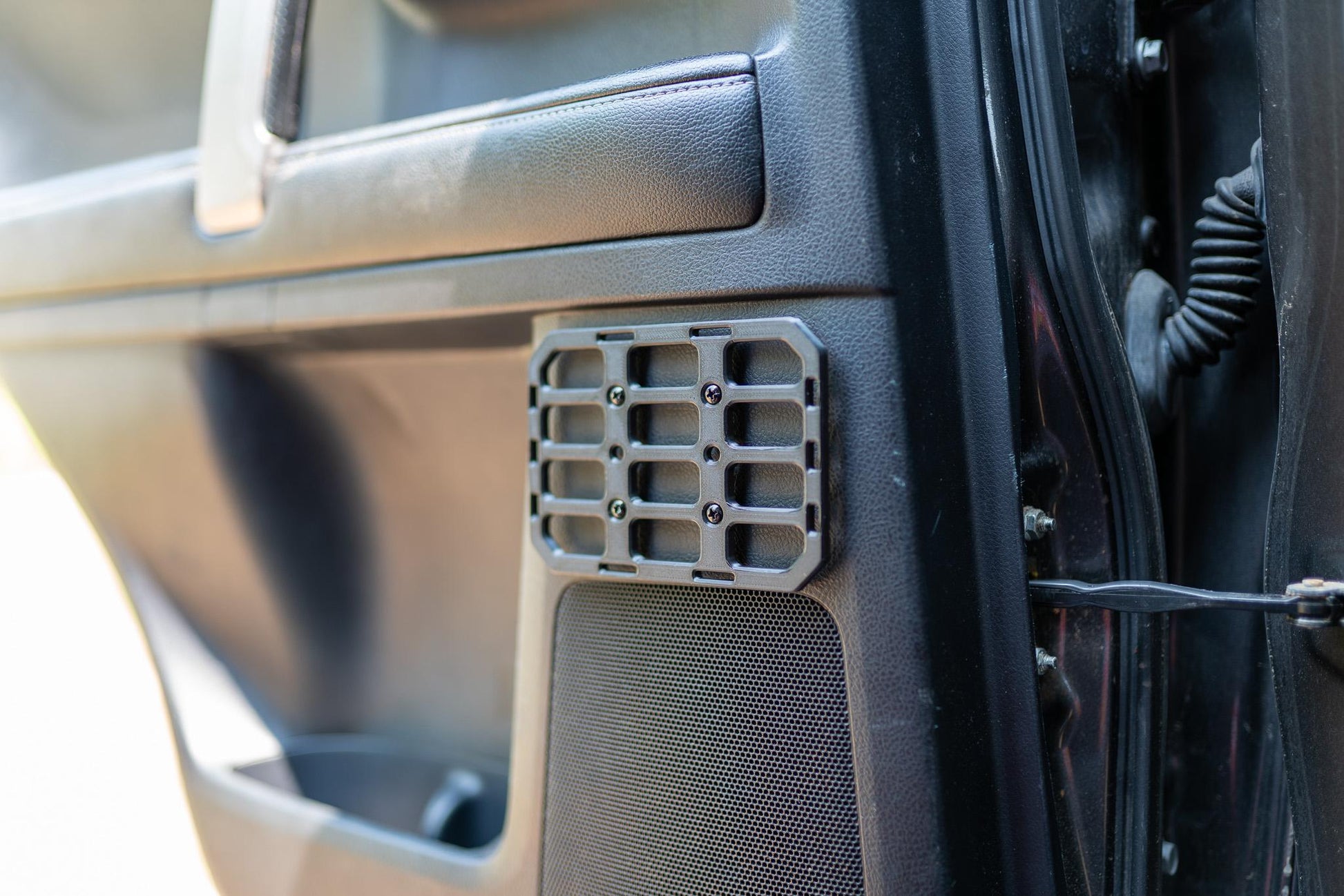 May include: A gray, rectangular MOLLE panel with a grid pattern is mounted on a car door panel. The panel is secured with black screws. The door panel is black and has a speaker and a door handle.