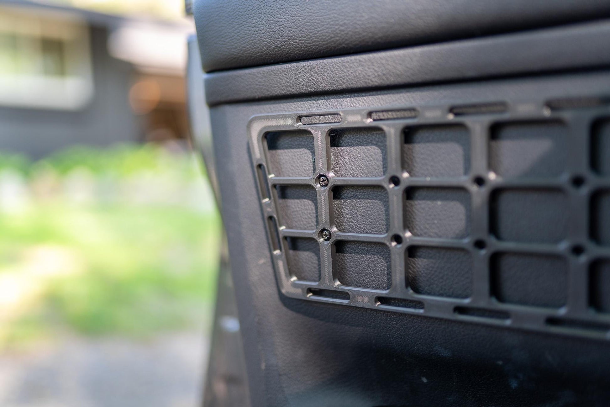 May include: A black, rectangular storage panel with a grid pattern, mounted on a vehicle's interior door panel. The grid is secured with small screws. The door panel has a textured surface.