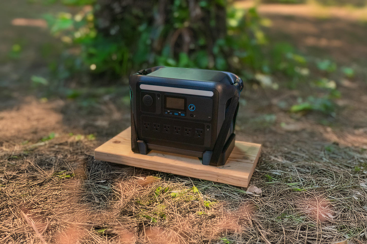 May include: A black portable power station with multiple outlets and a digital display, resting on a wooden platform. The power station is designed for outdoor use and is surrounded by grass and foliage.