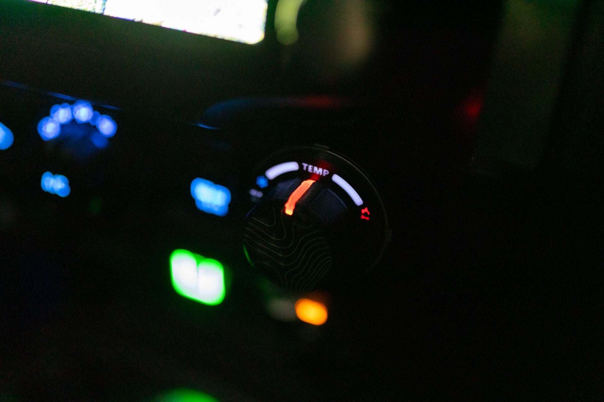 May include: Close-up of a car's dashboard with illuminated controls. The focus is on a temperature dial labeled "TEMP" with a red indicator. Other controls are visible with blue, green, and orange lights, suggesting a nighttime setting.