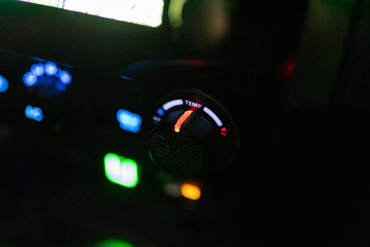 May include: Close-up of a car's dashboard with illuminated controls. The focus is on a temperature dial labeled "TEMP" with a red indicator. Other controls are visible with blue, green, and orange lights, suggesting a nighttime setting.